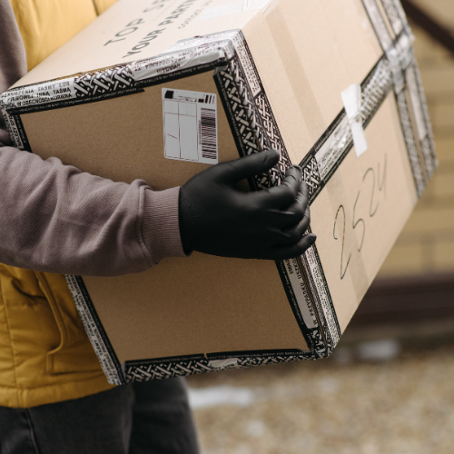 A person carrying a large cardboard package marked with delivery details, wearing gloves, symbolizing Amazon shopping or delivery services